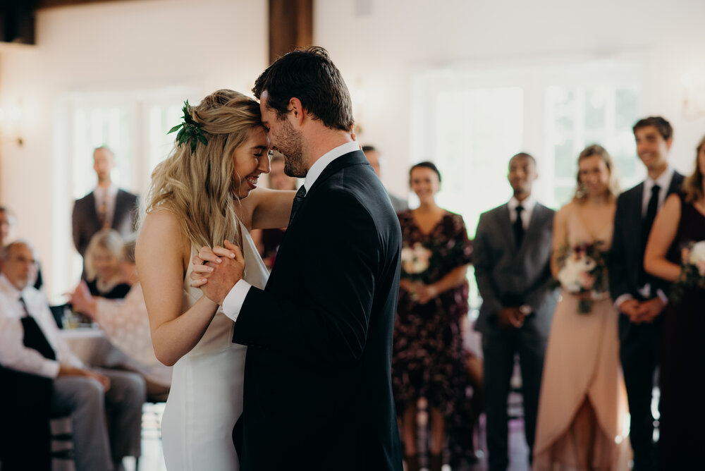 A bride and groom share a tender first dance at their indoor wedding reception, with the bride wearing a white gown and the groom in a black suit, surrounded by smiling guests in the background.