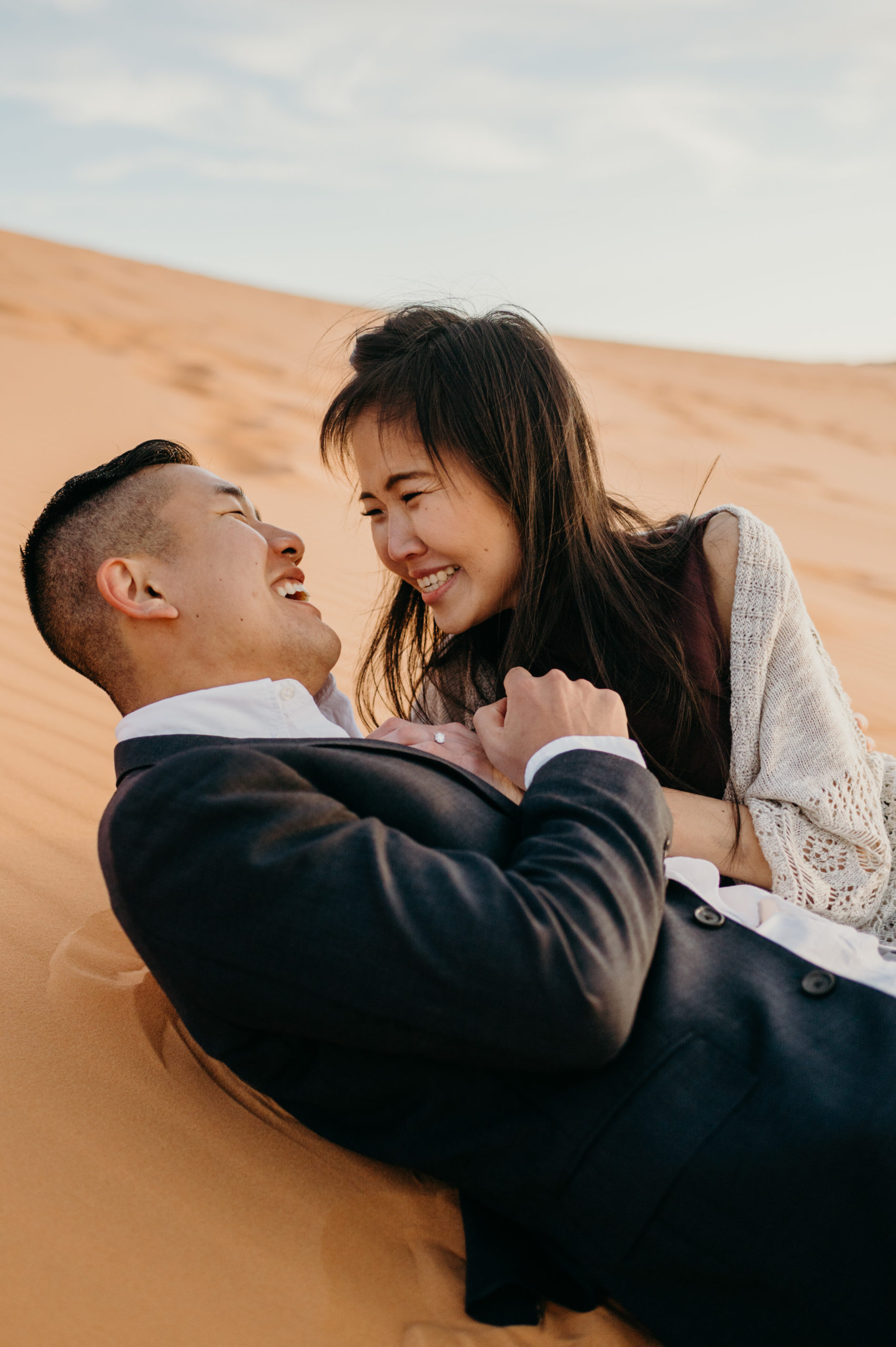 A joyful photo of a couple lying on the sand in a desert, smiling and holding hands, with the man wearing a suit and the woman in a light cardigan.