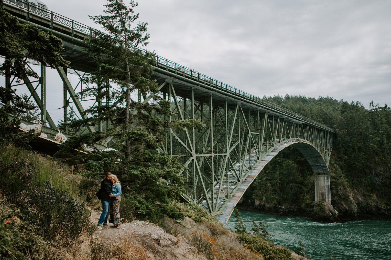 A scenic engagement photo of a couple standing near the Deception Pass Bridge, surrounded by lush greenery and overlooking the water below.