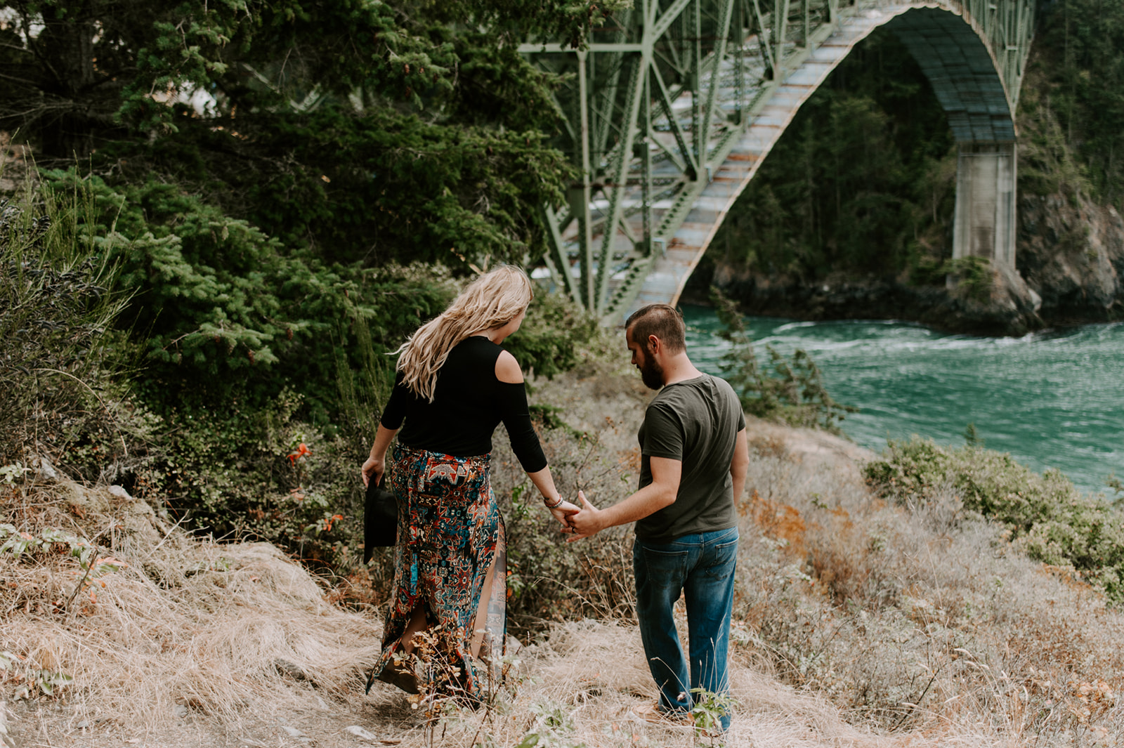 Couple walking hand in hand near a green steel bridge with a view of the water.