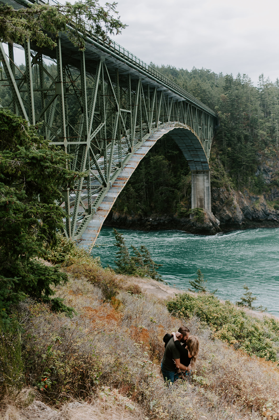 Couple sharing a kiss in a grassy area below a large steel bridge.