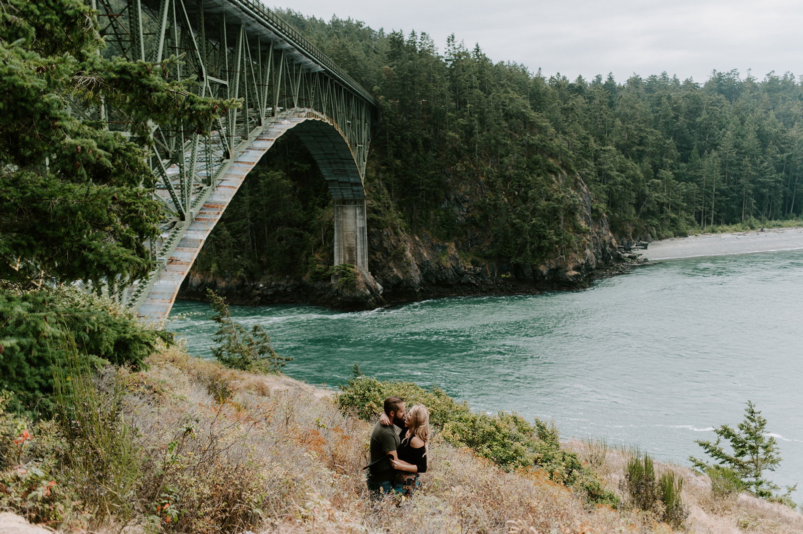 Couple sharing a kiss under a large metal bridge with the ocean in the background.