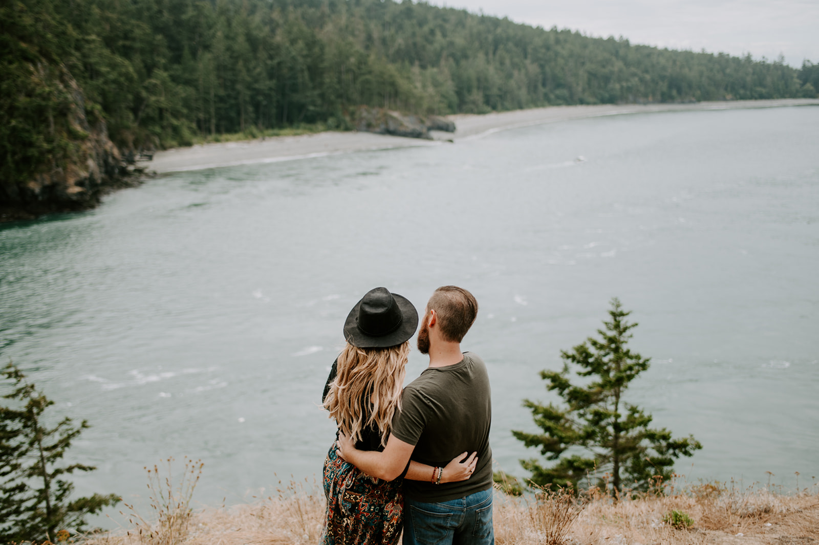 Couple embraces, standing on a cliff, looking out at the water with trees surrounding the beach below.