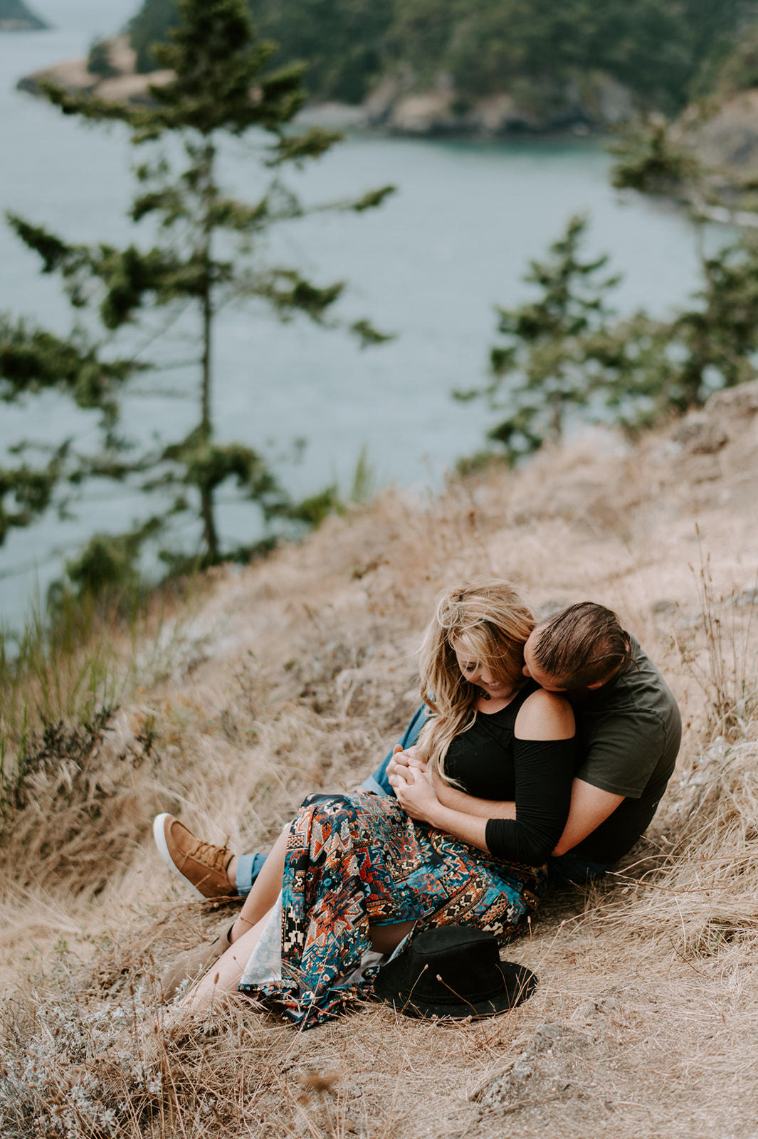 Couple sitting on a grassy hill, embracing and smiling with a serene view of the ocean in the background.