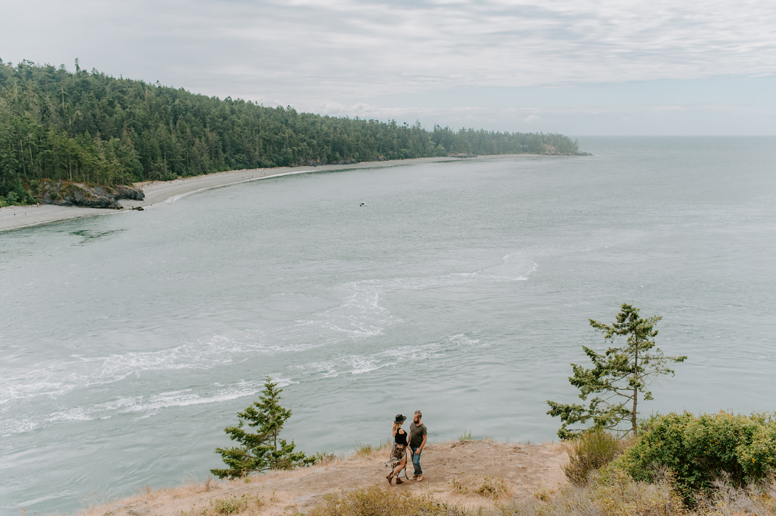 Couple walking on a cliffside overlooking a large bay with trees along the shoreline.