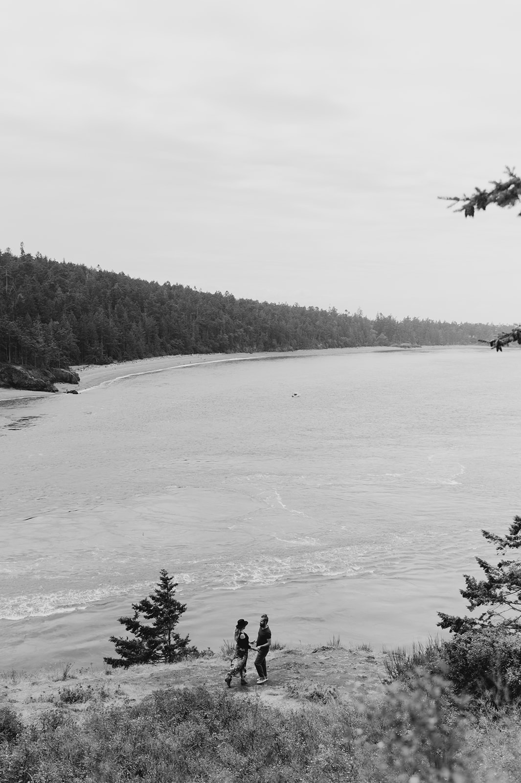 A black-and-white view of the couple walking along a cliffside, overlooking a large body of water.