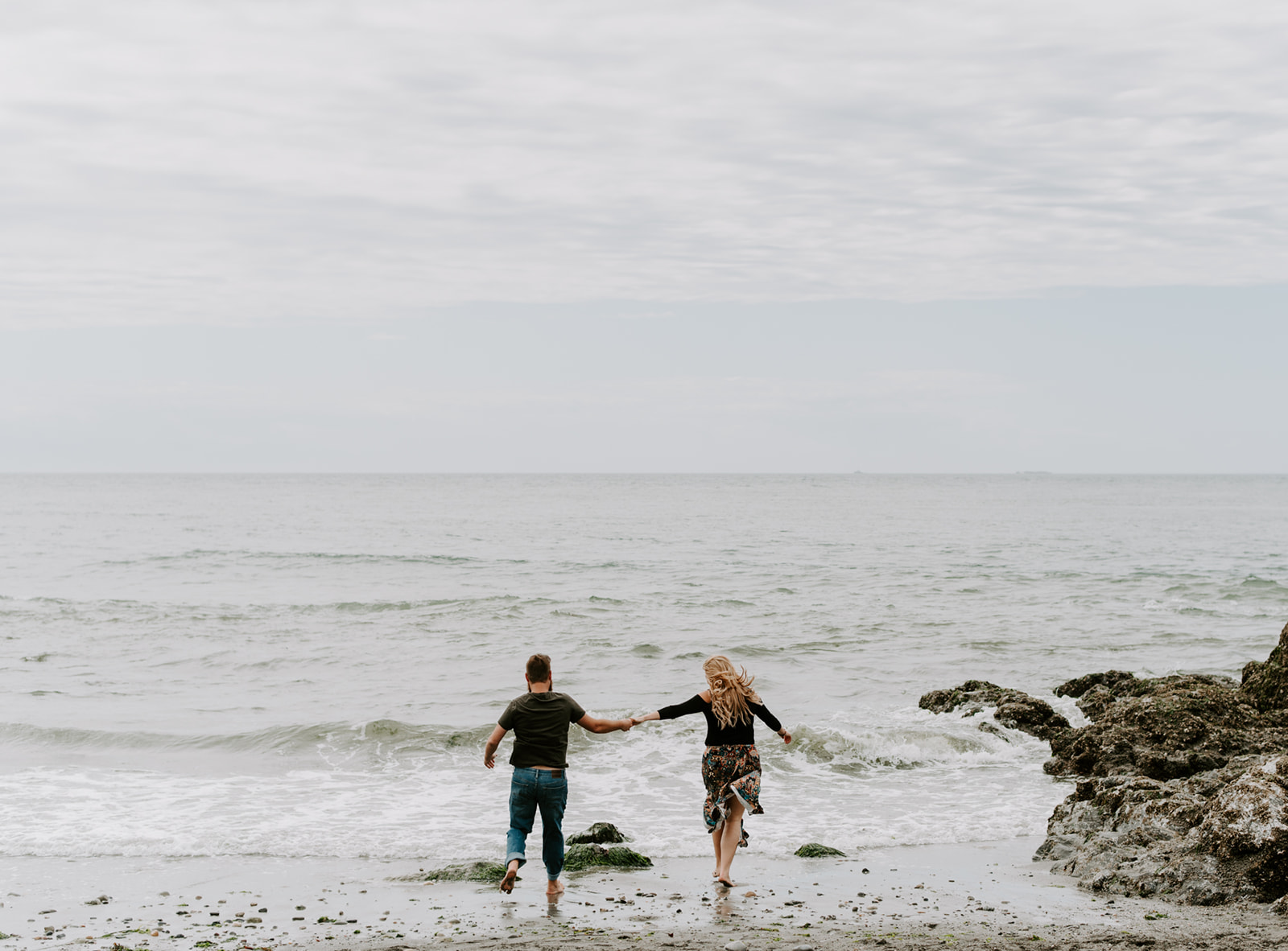 Couple running hand-in-hand toward the ocean during their Deception Pass anniversary session.