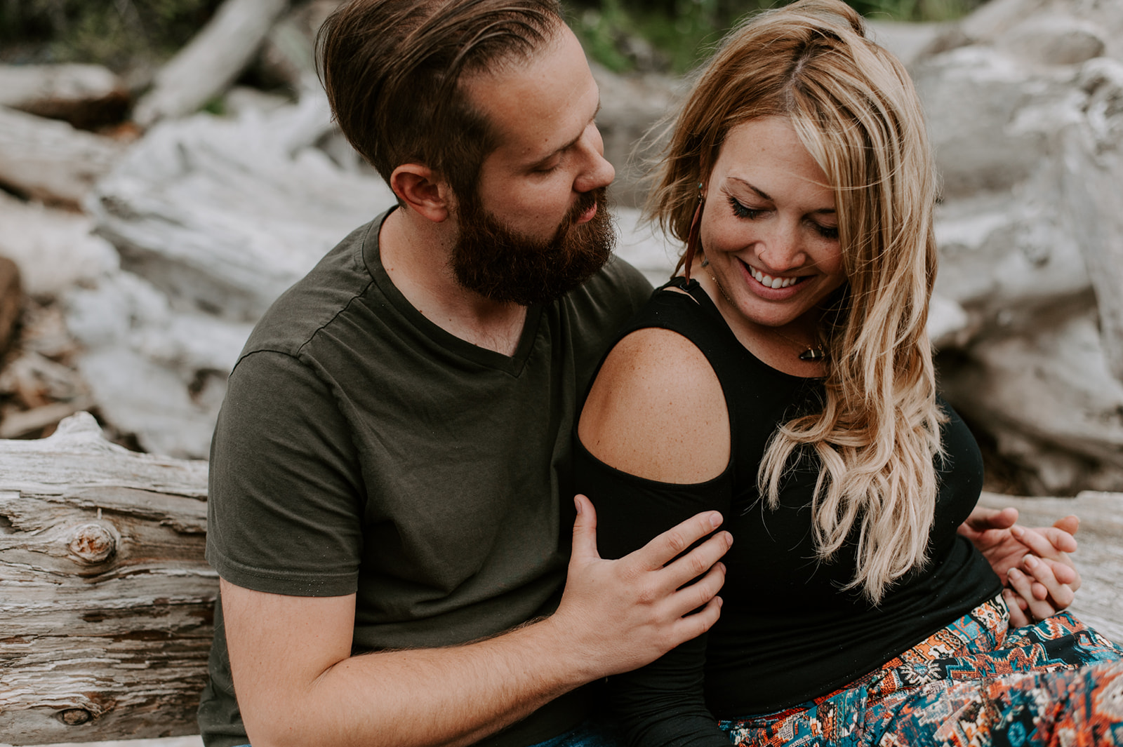 Couple sharing smiles and laughter while sitting together on driftwood during their Deception Pass anniversary session.