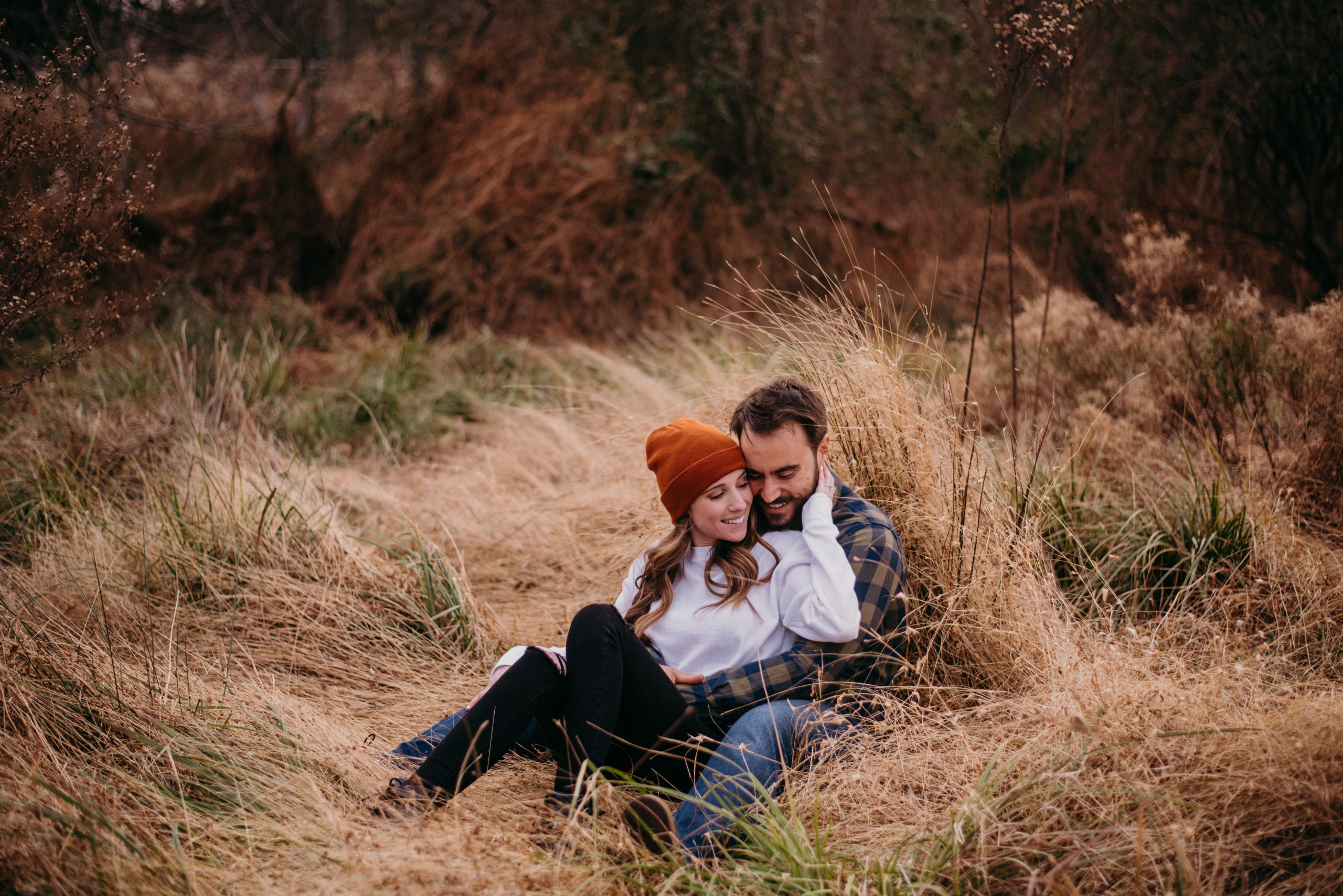 A cozy outdoor engagement photo of a couple sitting together in a grassy field, the woman wearing a brown beanie and white sweater, leaning into the man who is wearing a plaid shirt.