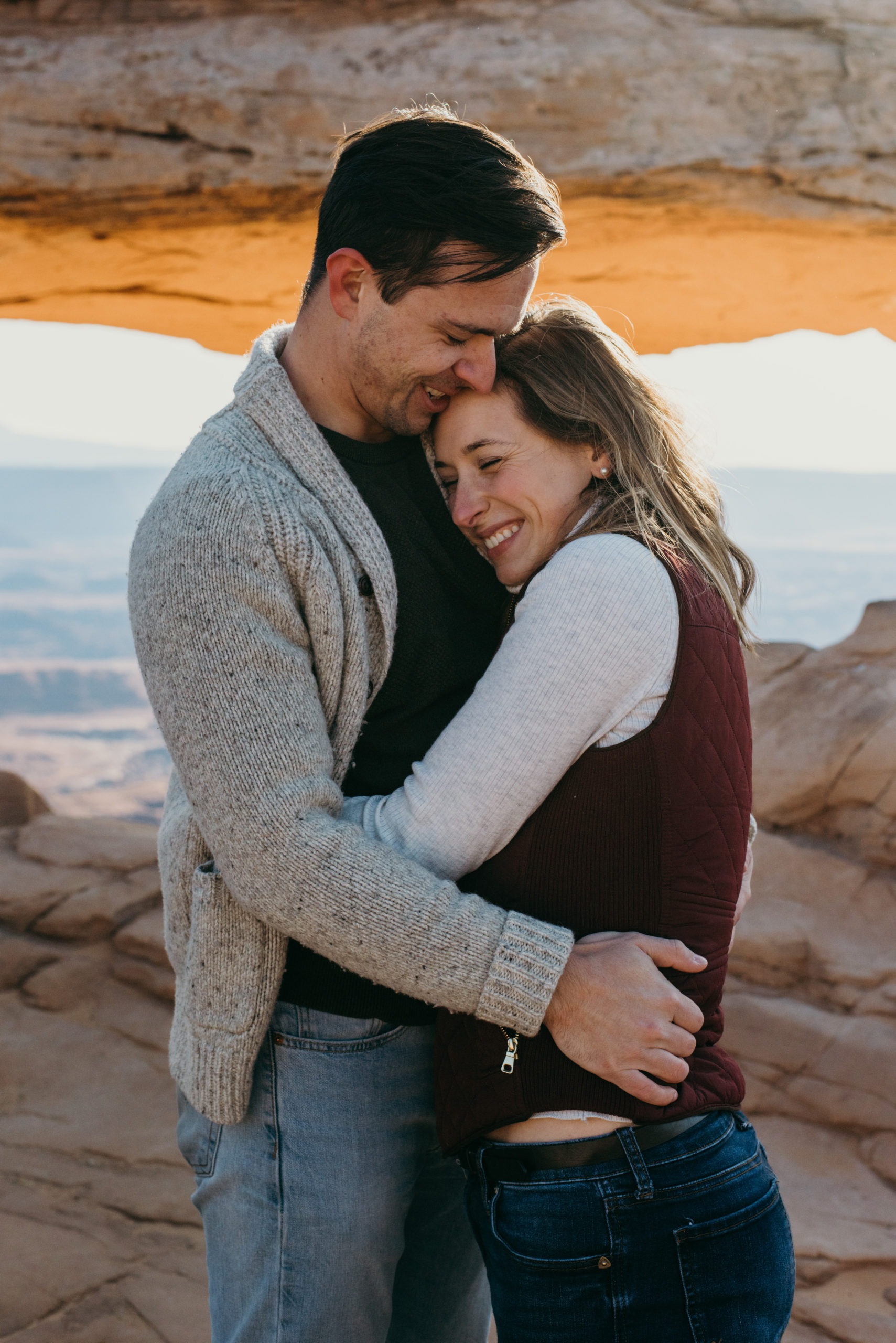 A couple sharing a tender embrace in a rocky landscape with a natural arch in the background, both smiling and enjoying the moment.