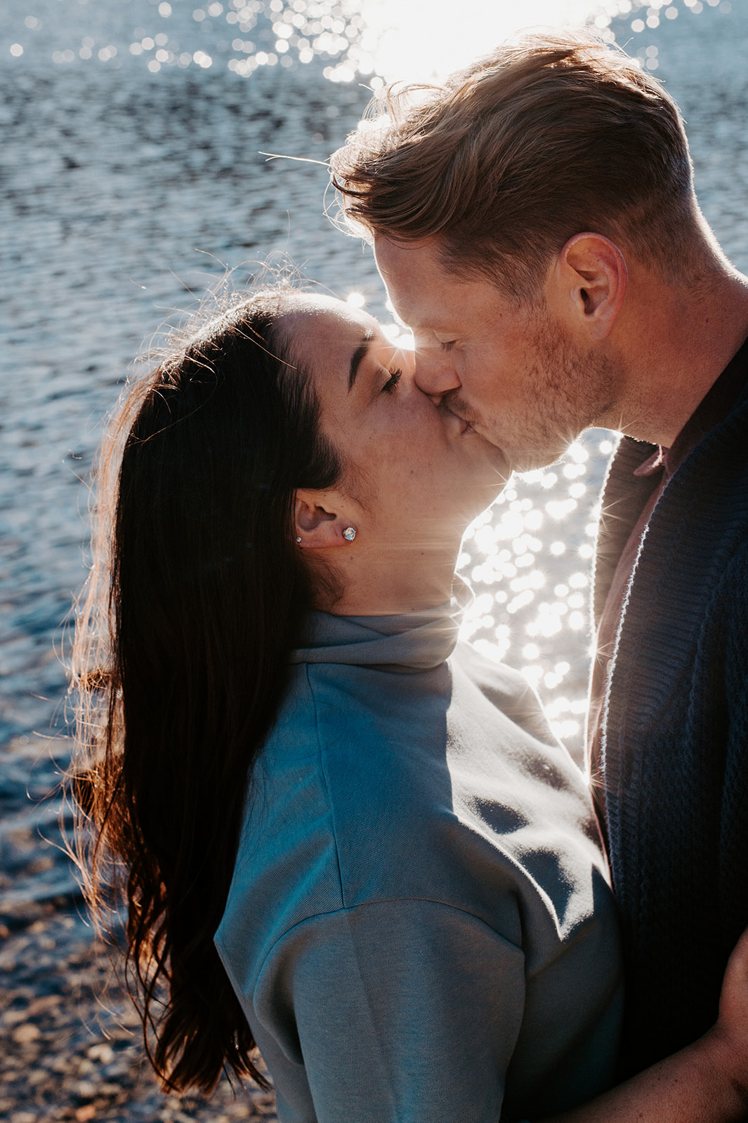 A couple sharing a kiss by the sparkling water, with sunlight reflecting off the surface and creating a romantic glow around them.