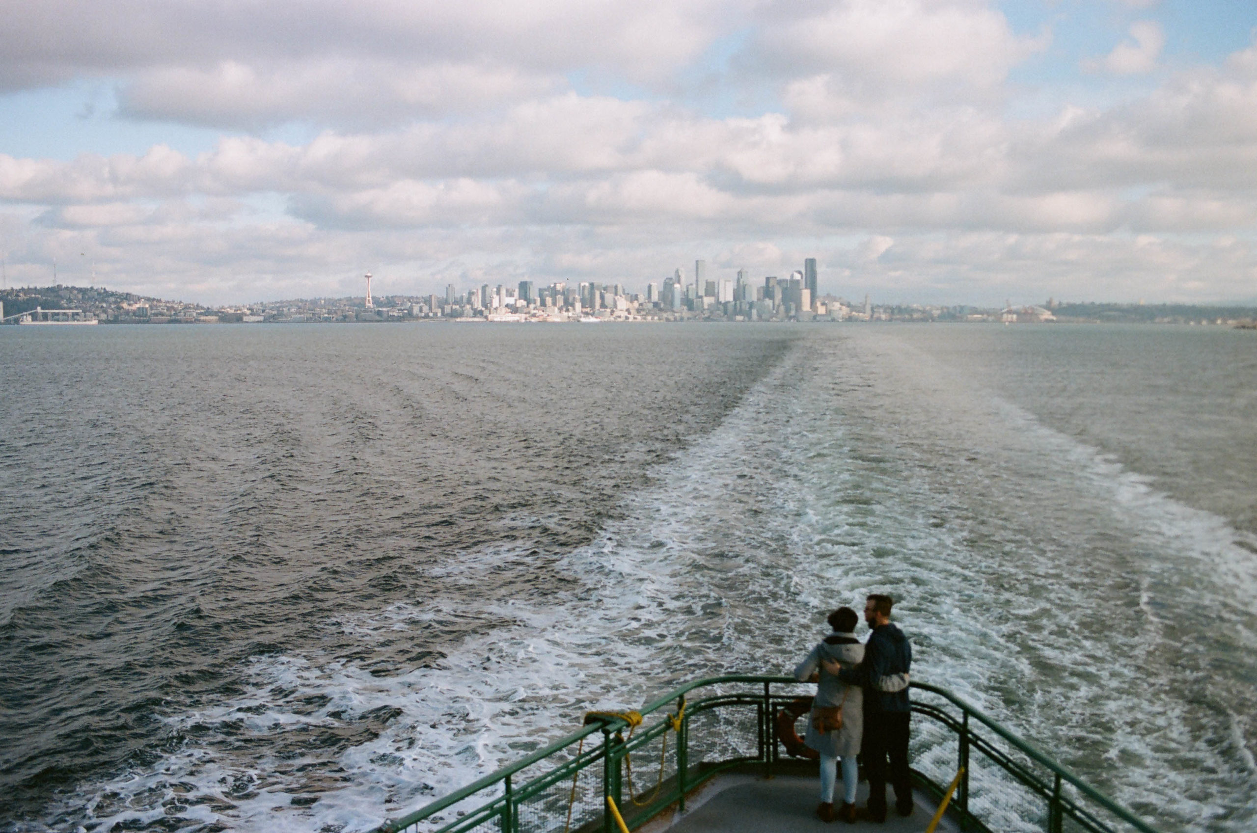 A couple embracing on the deck of the Washington Ferry with the Seattle skyline in the background, under a partly cloudy sky.