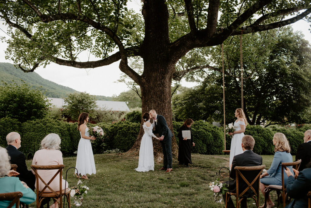 Couple kissing during a creative outdoor wedding ceremony under a large oak tree