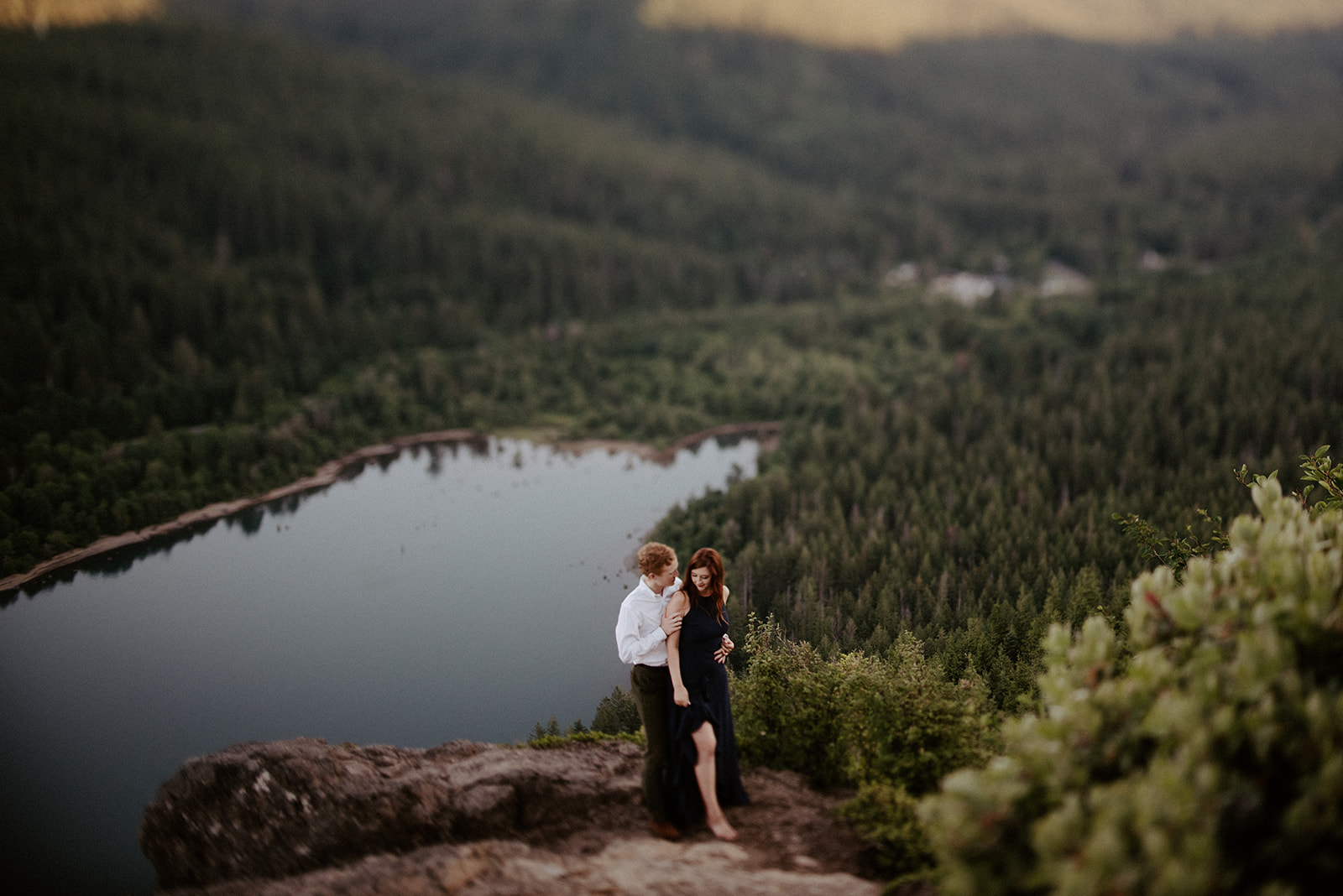 A couple standing on a cliff, surrounded by lush forest and overlooking a serene lake.