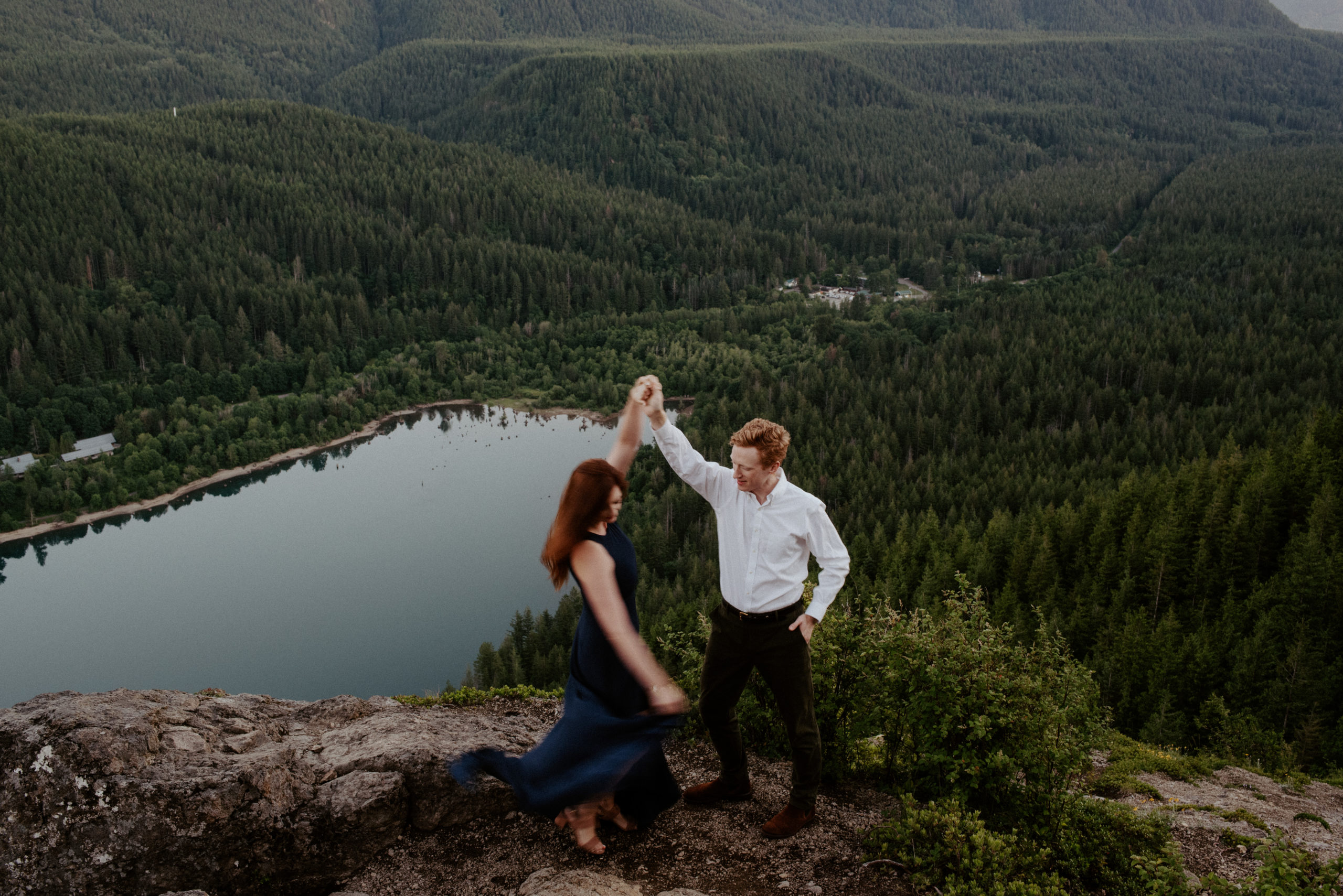 A couple dances on a mountaintop, with the man in a white shirt and the woman in a navy dress, surrounded by a stunning view of the forest and a lake below, capturing a romantic and adventurous moment.