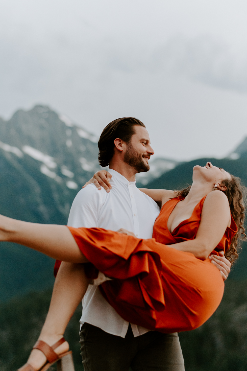 A couple dances outdoors with mountains in the background, the man wearing a white shirt and the woman in a vibrant orange dress, as they share a joyful and carefree moment.