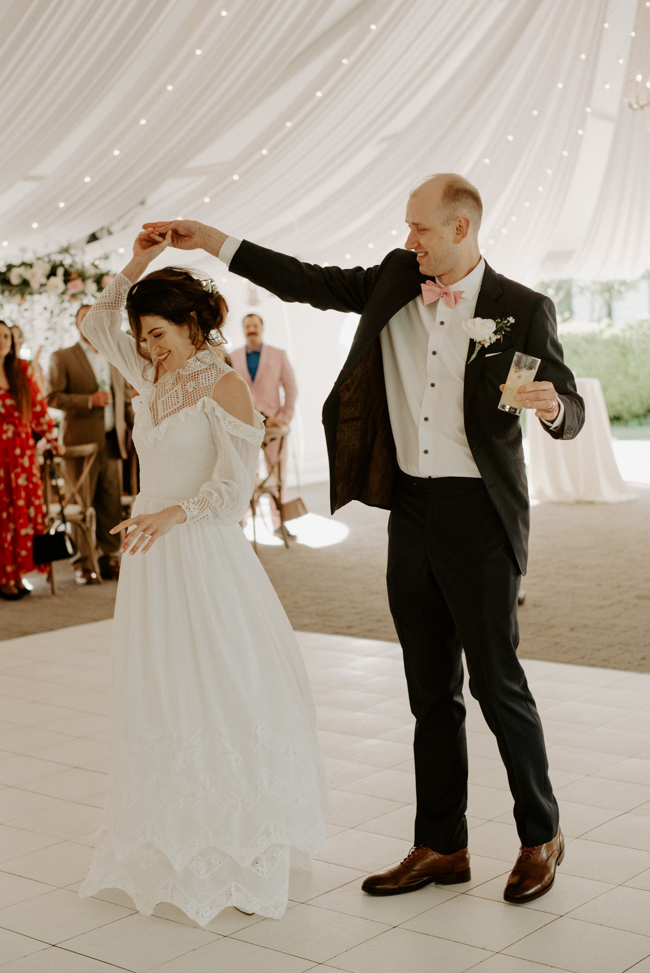 A bride and groom share their first dance under a white canopy at their wedding, with the groom twirling the bride while holding a drink, creating a joyful and elegant atmosphere with guests in the background.
