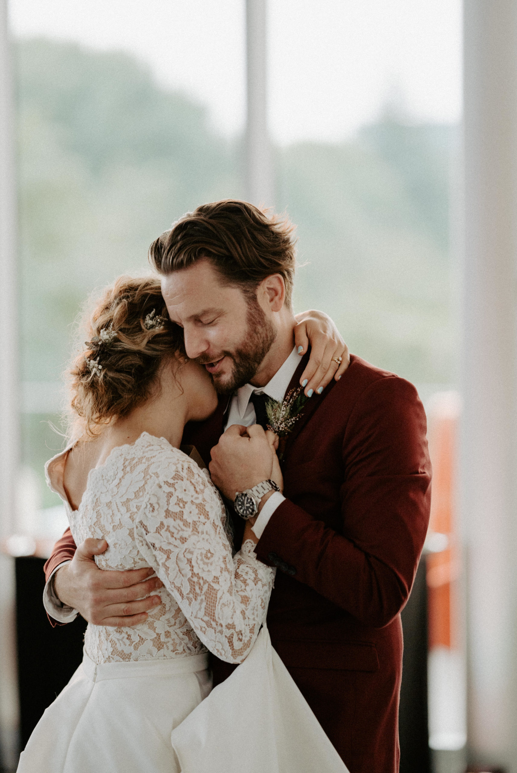 A bride and groom share an intimate embrace during their first dance at their wedding, with the bride wearing a lace gown and the groom in a burgundy suit, their foreheads touching and eyes closed, capturing a tender and emotional moment.