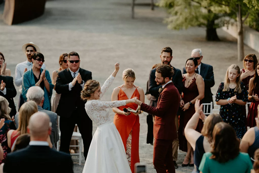 Bride in white lace dress and groom in maroon suit celebrate with raised hands among cheering wedding guests.