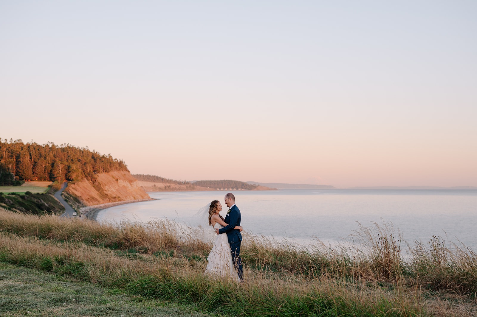 Bride and groom embracing in tall grass on the bluffs at Ebey's Landing at sunset, with the Strait of Juan de Fuca and cliffside coastline stretching behind them, Whidbey Island Washington
