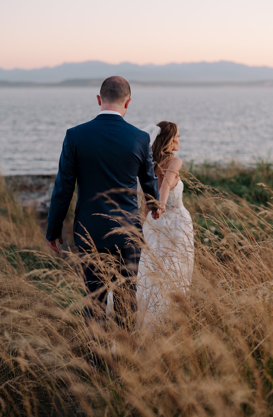 Bride and groom walking hand in hand through tall golden grass at Ebey's Landing with the Strait of Juan de Fuca and Olympic Mountains visible at sunset, Whidbey Island Washington