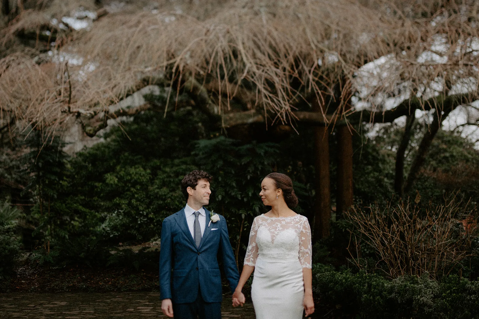 A bride and groom in wedding attire smiling at each other while holding hands in Parsons Gardens.