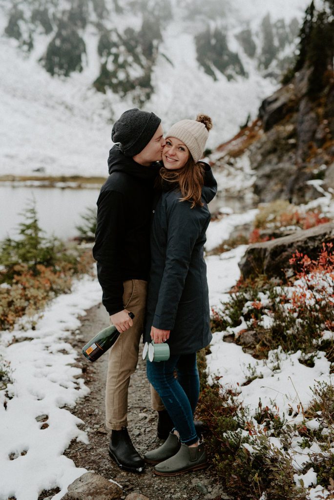 men kissing his fiancé's cheek both wearing winter's outfit at a snow covered hill area
