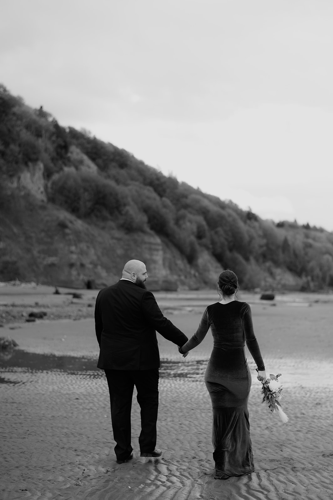 Bride and groom holding hands while walking along the sand with forested cliffs in the background.