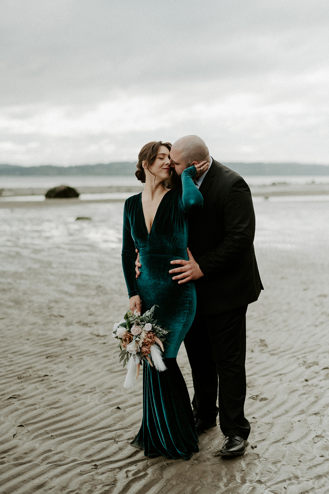 A groom embraces his bride on a beach, her bouquet featuring muted florals and pampas grass.