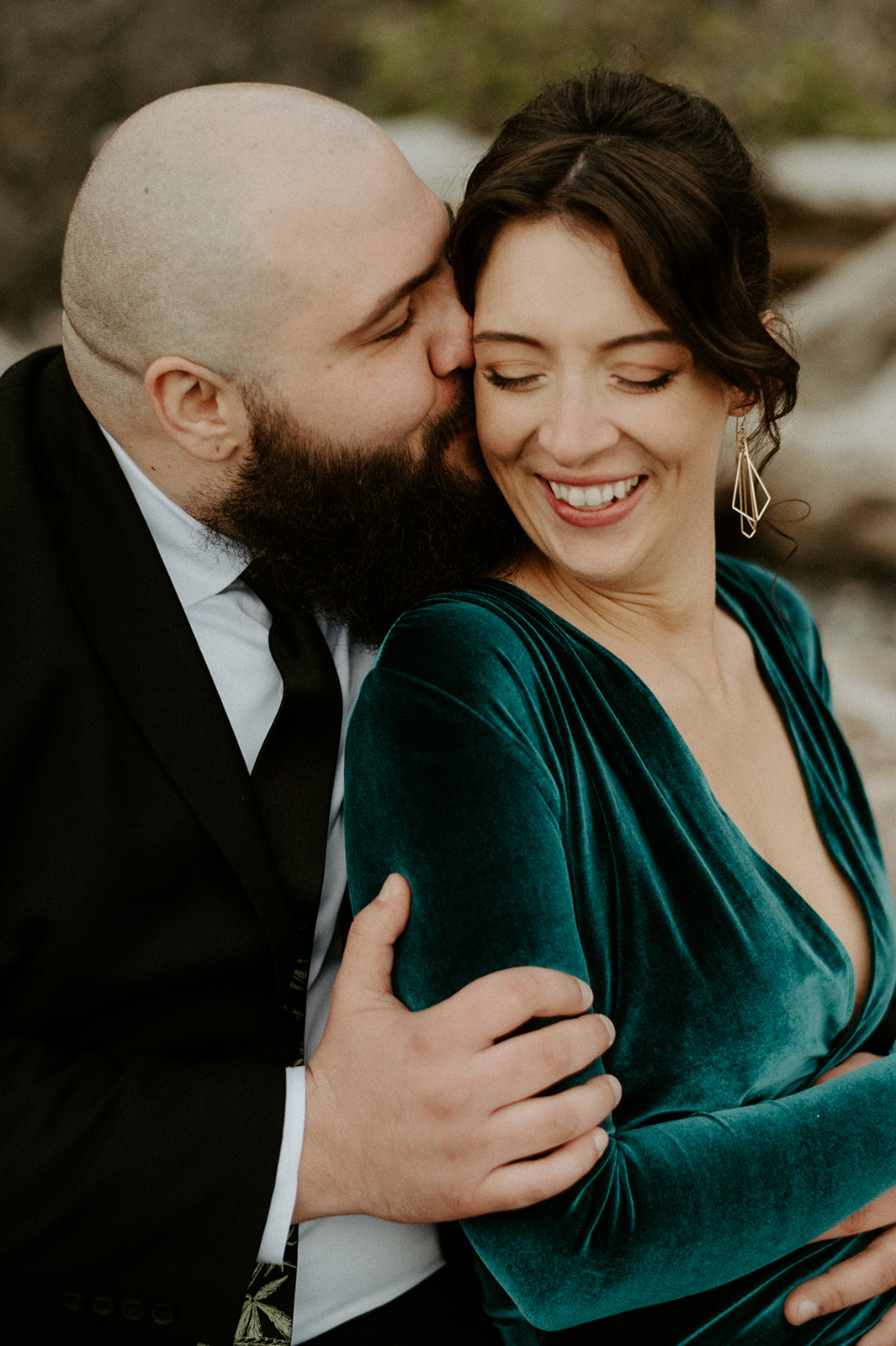 A groom kisses his smiling bride's cheek as she laughs, wearing an emerald green velvet dress.