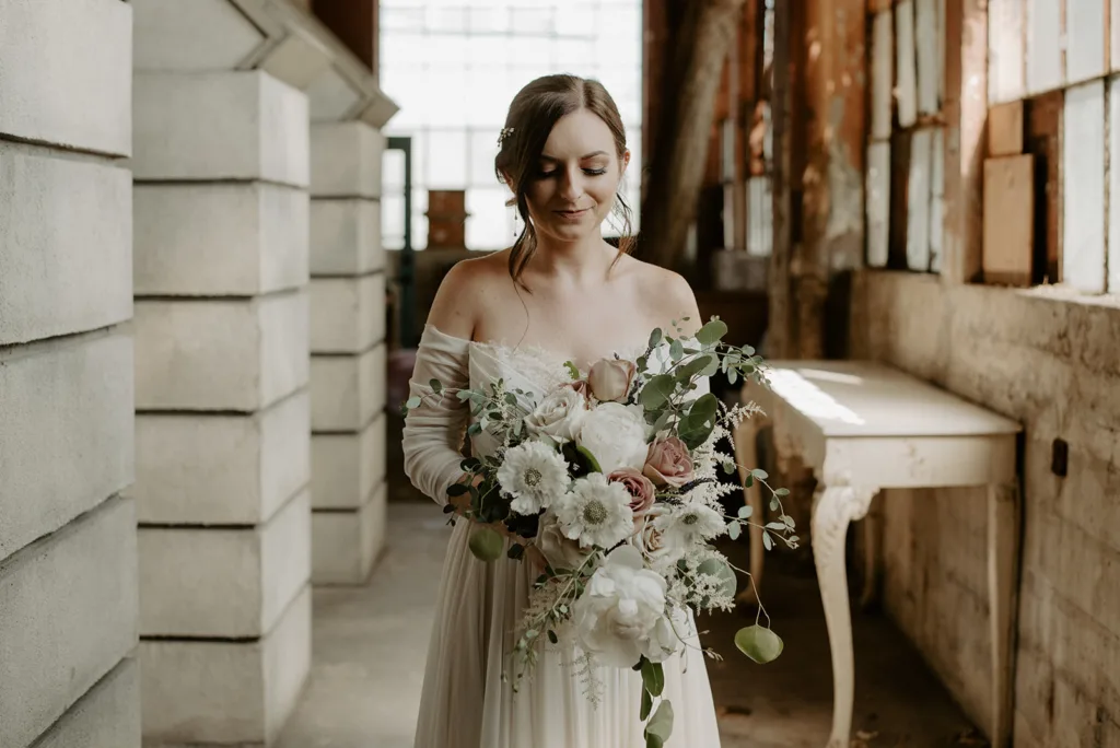 Bride holding a floral bouquet in a rustic setting with exposed brick walls and soft natural light.
