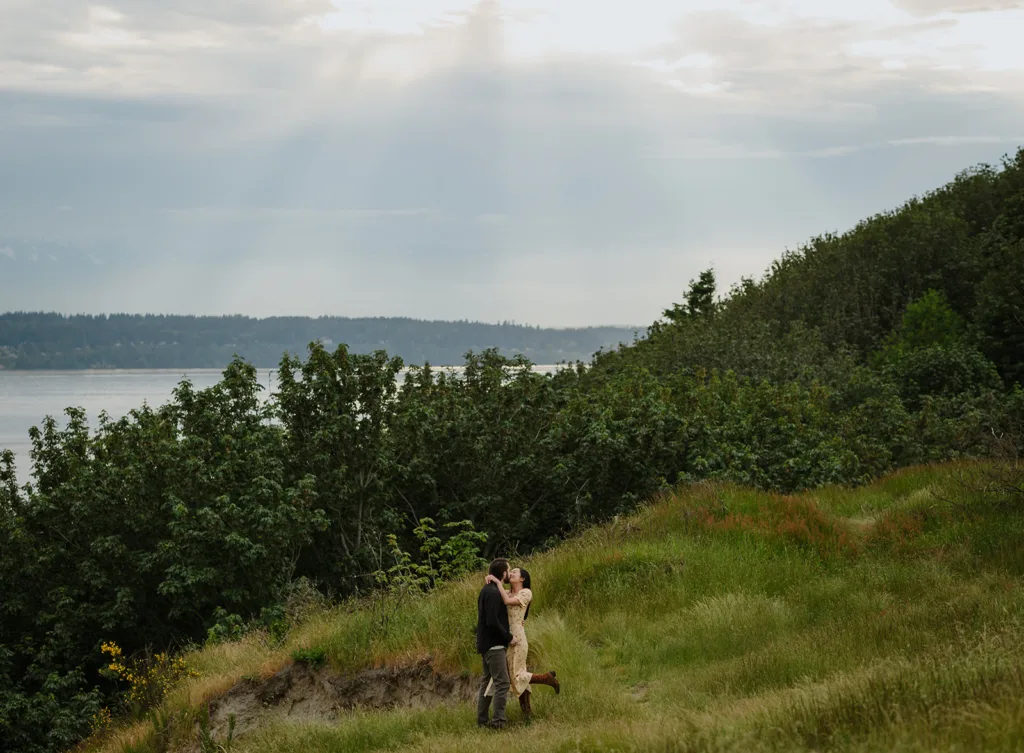 A picturesque moment of a couple embracing on a grassy hillside, with lush greenery and a serene body of water in the background under a cloudy sky.