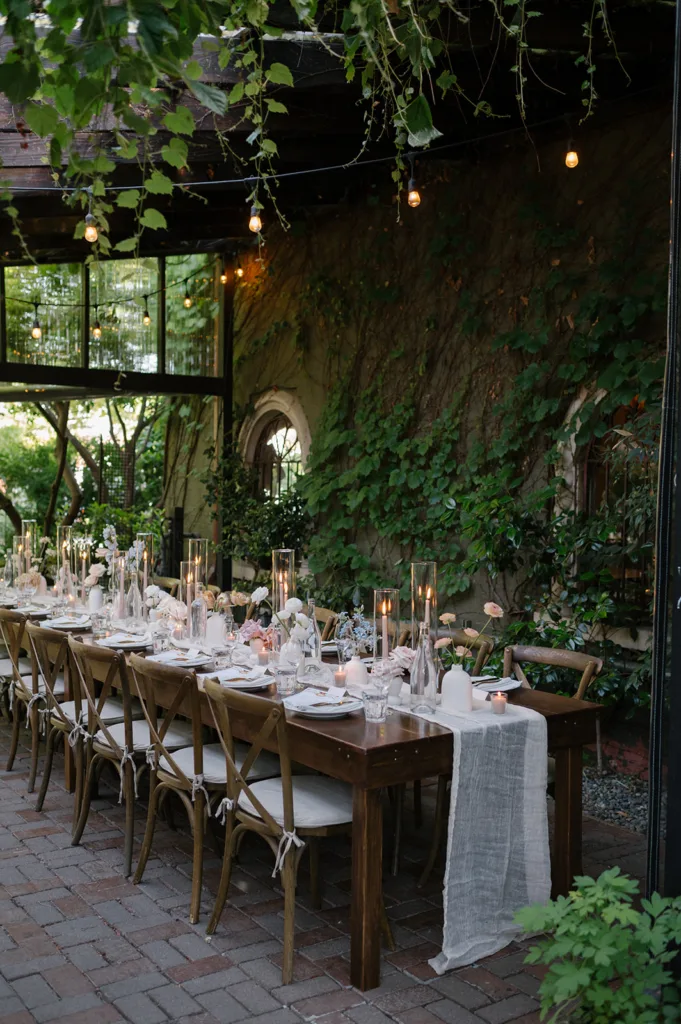 Elegant outdoor wedding reception setup at The Corson Building with rustic wooden tables, delicate floral arrangements, and warm string lights, encircled by ivy-clad walls for a romantic Seattle evening.