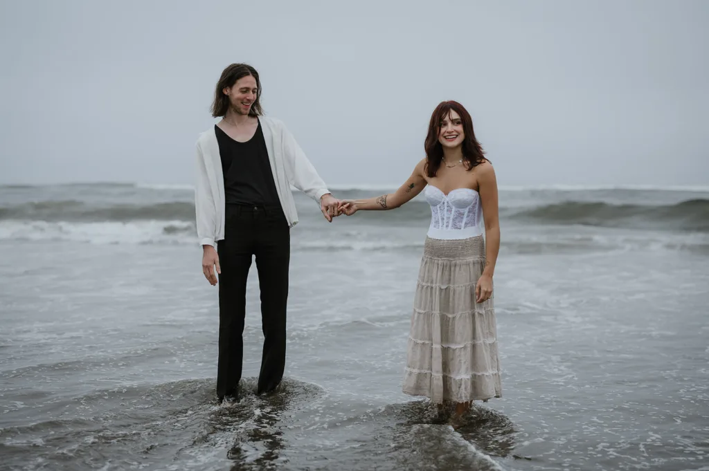 Engaged couple playfully holding hands in the ocean waves on the Oregon coast