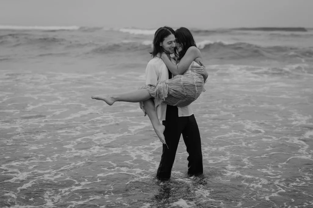 Man carrying woman in his arms on the beach in a black and white Oregon coast engagement photo