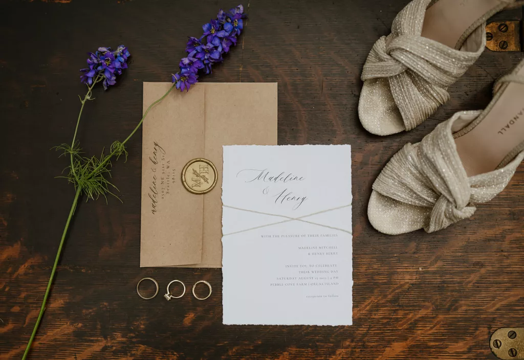 A flat lay of wedding details showcasing elegant invitations with calligraphy, a wax seal, sparkling bridal shoes, and rings alongside a sprig of purple flowers for a touch of natural beauty.