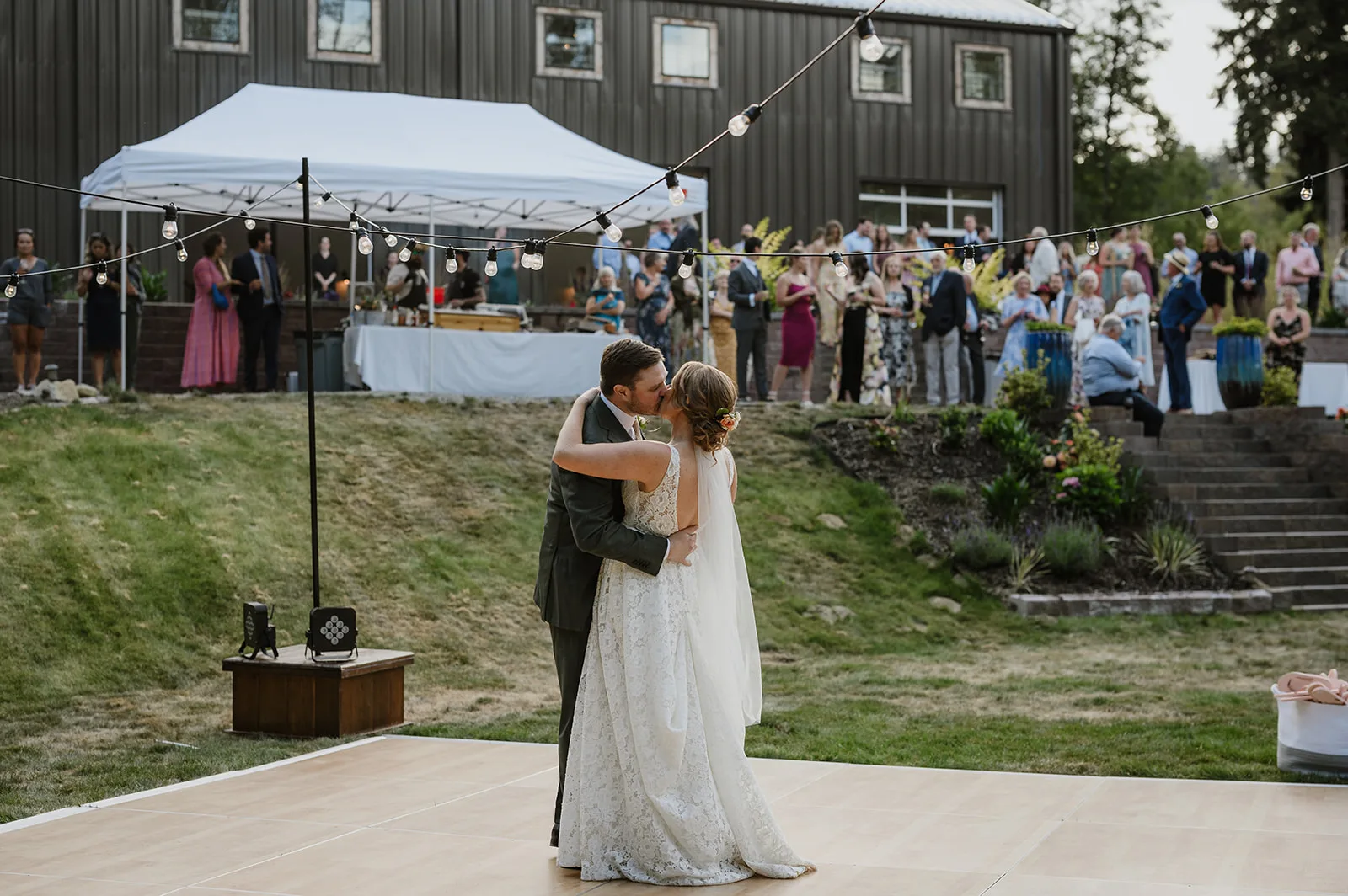 A bride and groom share their first dance under string lights at an outdoor wedding, with the bride wearing a lace gown and the groom in a dark suit, surrounded by guests who are watching and celebrating in the background.