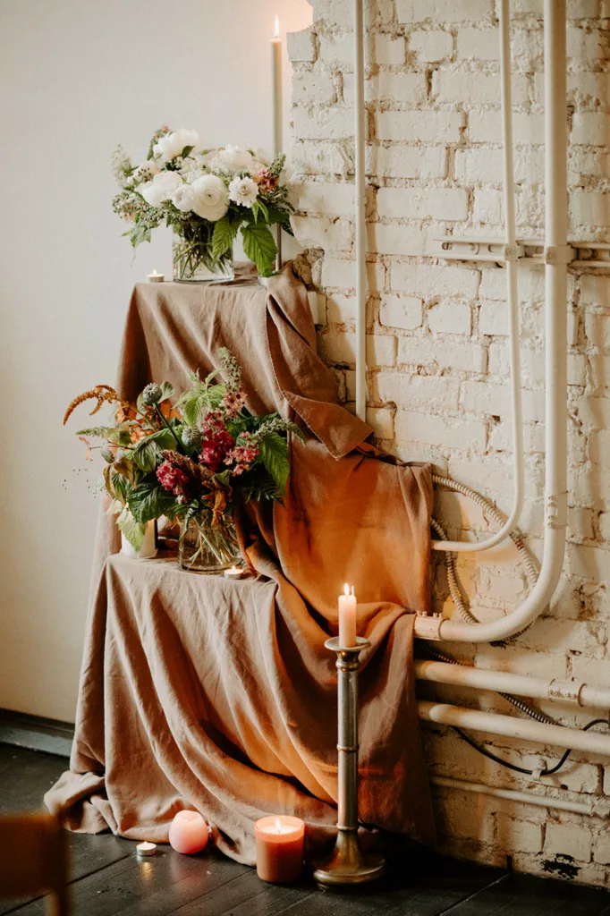 A rustic chic wedding decor corner with earth-toned drapery, vibrant floral centerpiece, and glowing candles set against a white brick backdrop for a touch of DIY sophistication.