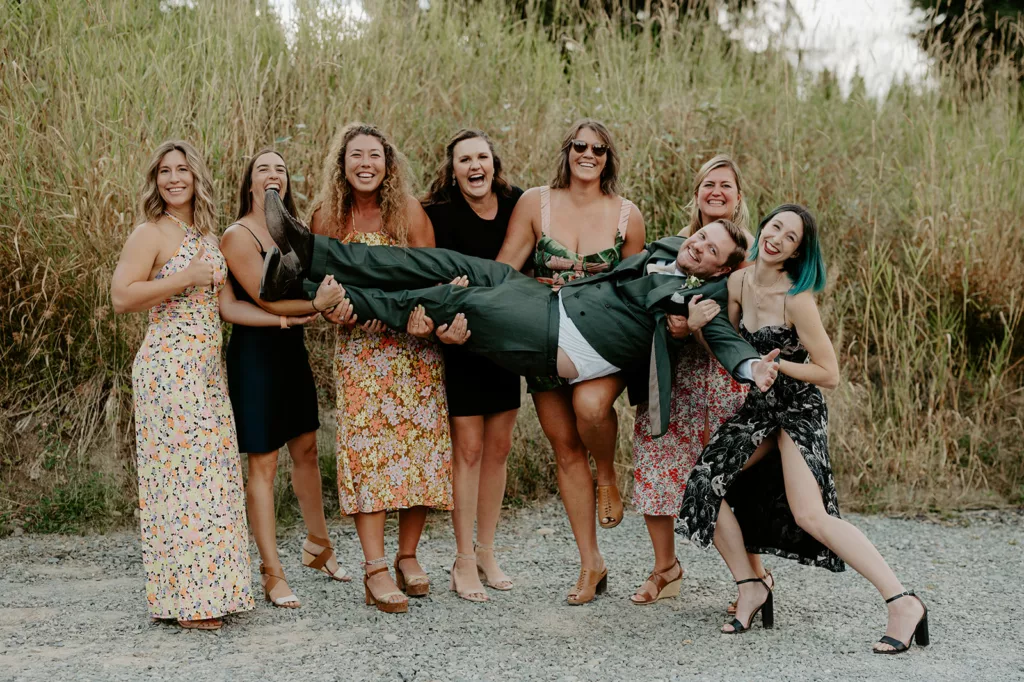 A group of seven women in summer dresses joyfully lifting a smiling groom in a green suit against a backdrop of tall reeds.