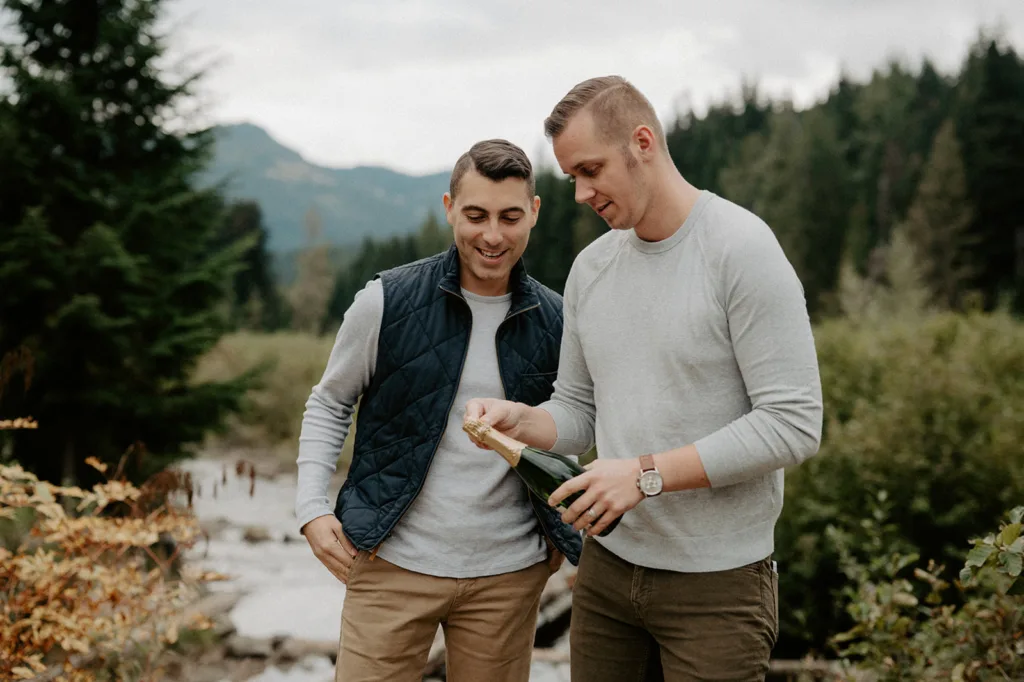 Two smiling men in casual attire open a bottle of champagne in a forested area, with a gentle river and rolling hills in the background.