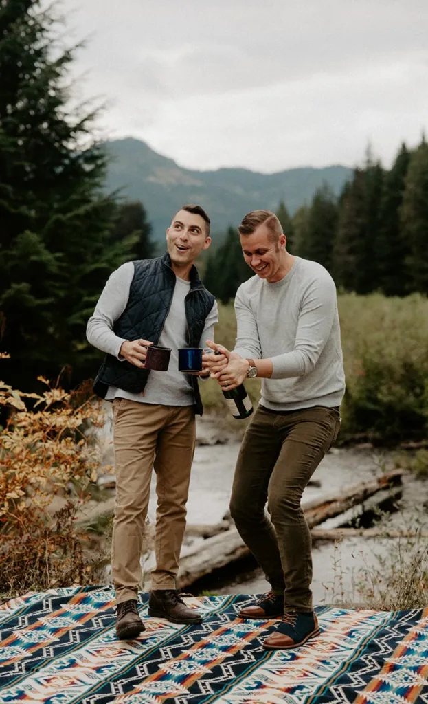 "Two men sharing a joyful moment, opening a bottle of wine on a patterned blanket in a forest clearing, with a tranquil river and mountains in the backdrop."