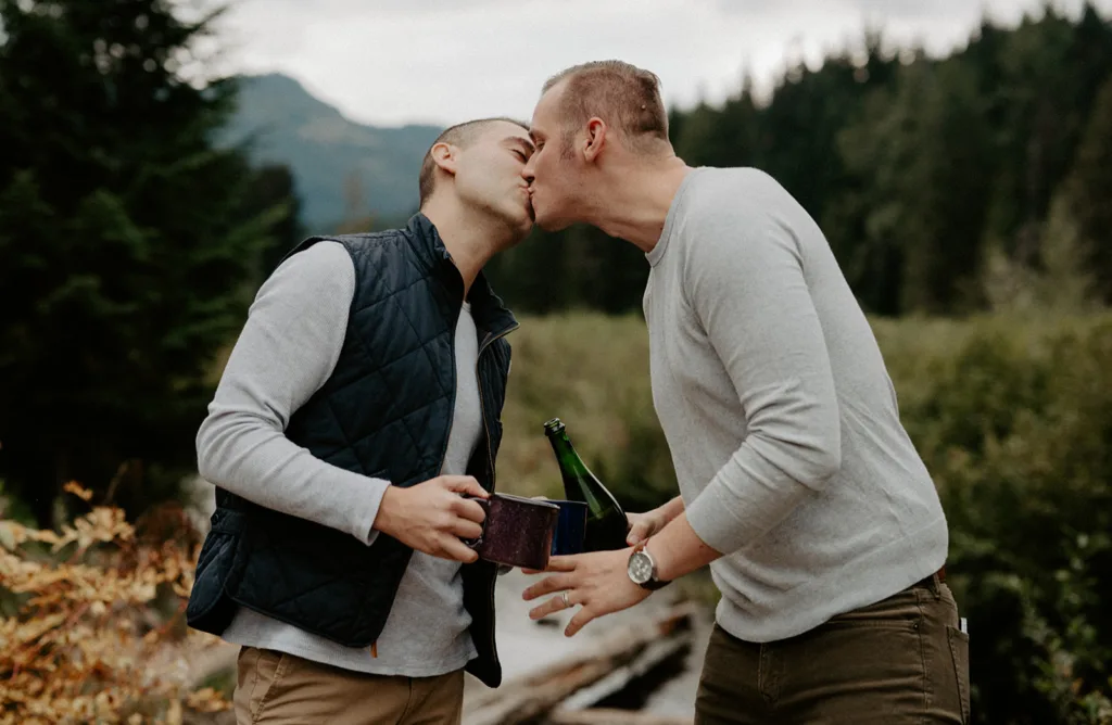 "Two men sharing an intimate kiss, holding a bottle of champagne, with a lush forest backdrop."