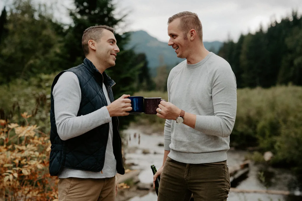 "Two men, sharing a toast with enamel mugs in a forest, smile at each other, with a river and mountains in the background."