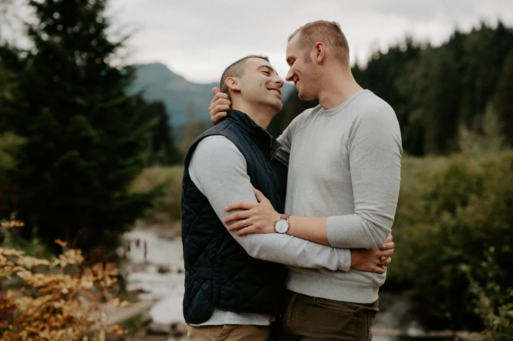 "Two men in a warm embrace, smiling at each other with a backdrop of lush trees and a mountain vista."