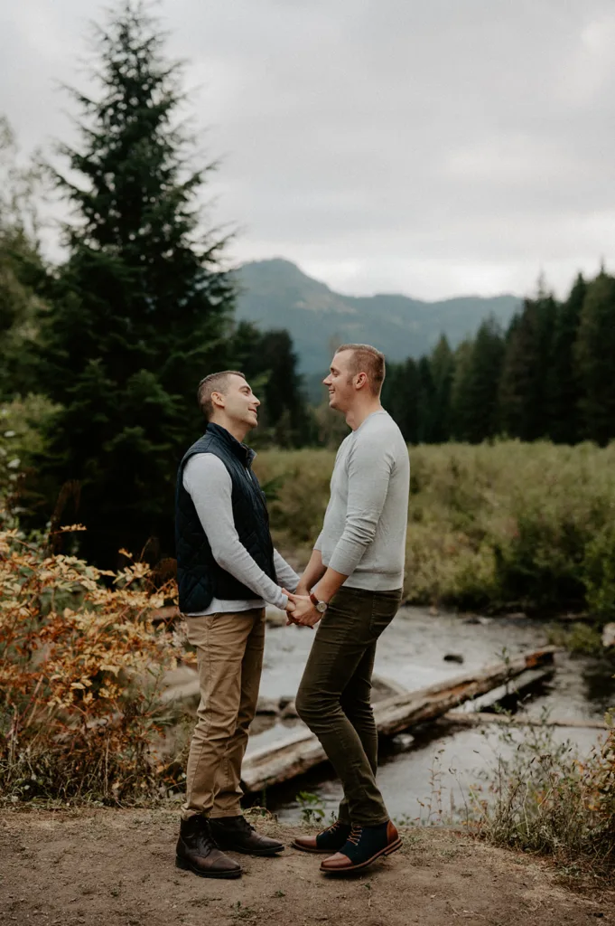 Two men holding hands, standing face to face with genuine smiles amidst a serene natural backdrop.