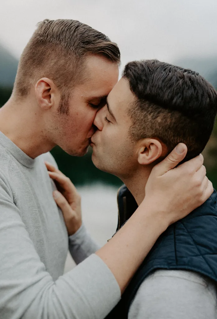 Close-up of two men sharing a tender kiss, hands gently cradling each other's face and shoulder against a blurred natural backdrop.