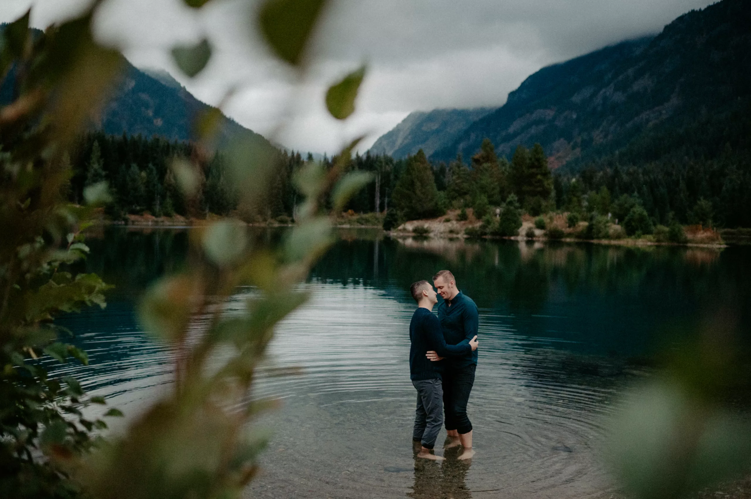 A romantic moment of a couple standing barefoot in a mountain lake, surrounded by forested hills and misty mountains.