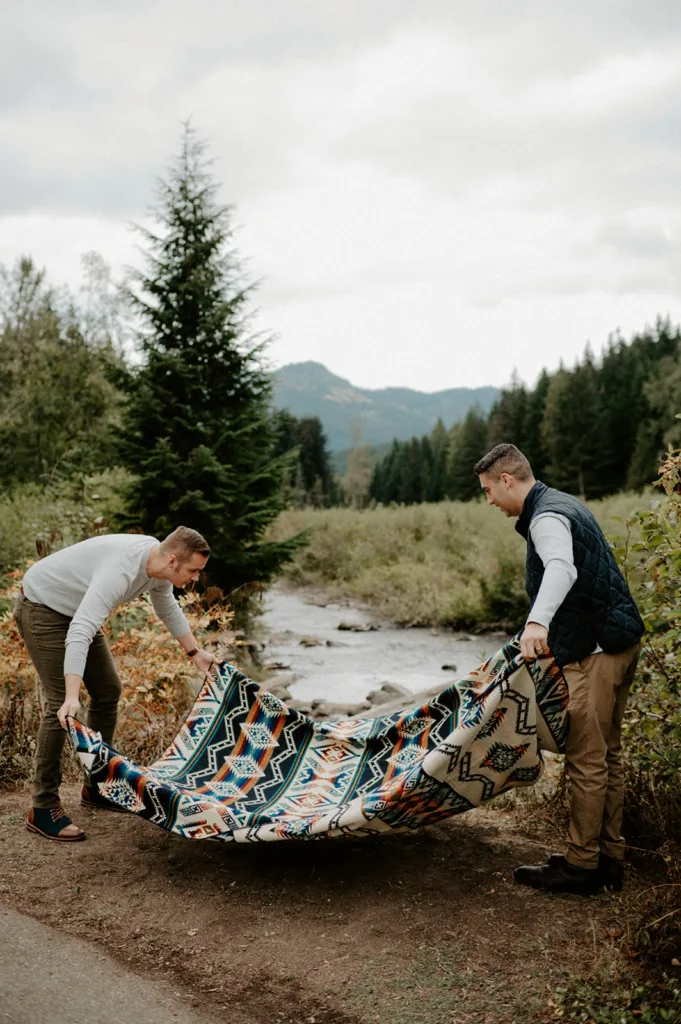 Two men partake in a playful moment while spreading a vibrant, patterned blanket beside a serene creek with a backdrop of lush coniferous trees and distant mountains under an overcast sky.