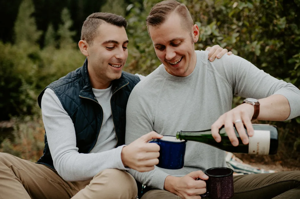 "Two men sharing a lighthearted moment as they pour a bottle of champagne into mugs outdoors."