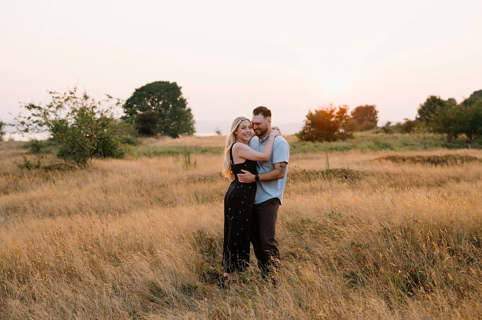 A couple embracing in a sunlit meadow with tall grass, trees in the background, and a warm sunset creating a romantic atmosphere.